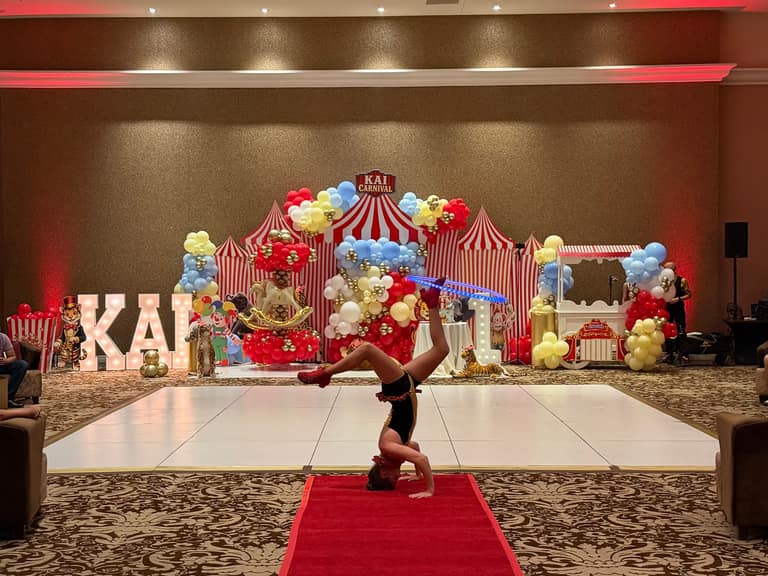 A circus-themed party installation with striped tents, balloon garlands, a marquee, and a performer on a red carpet