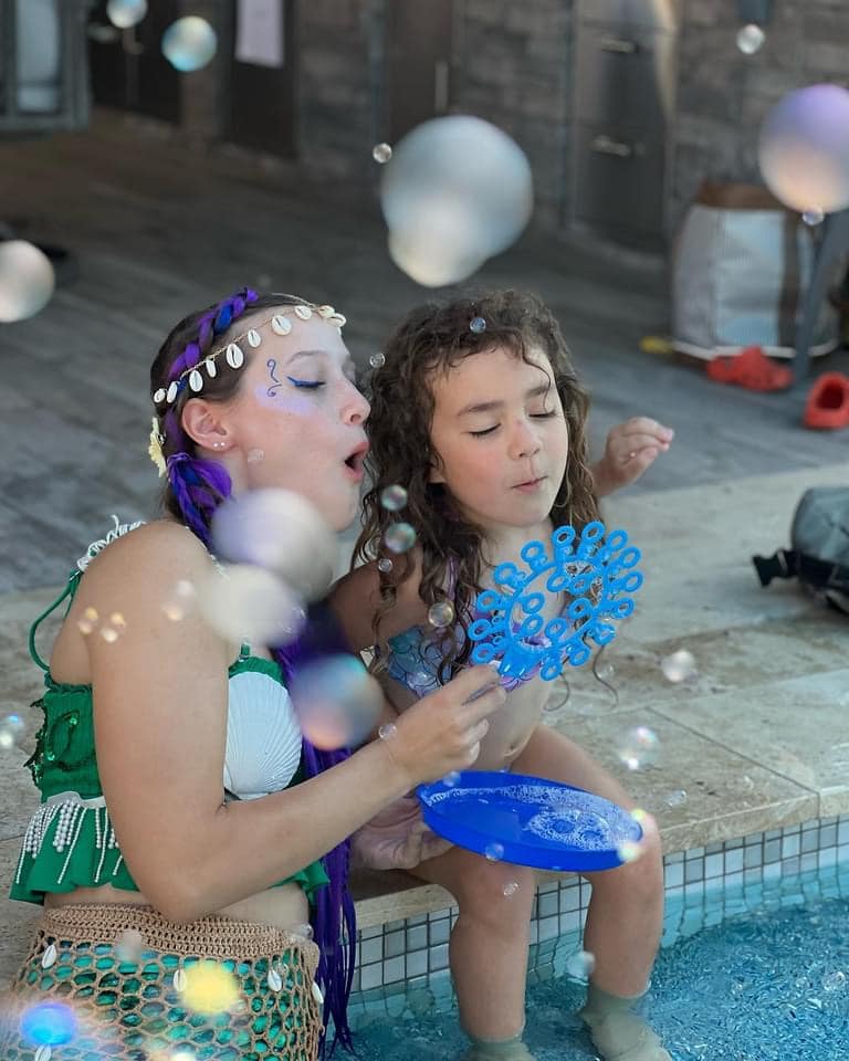 A mermaid performer blows bubbles with a child at a poolside party