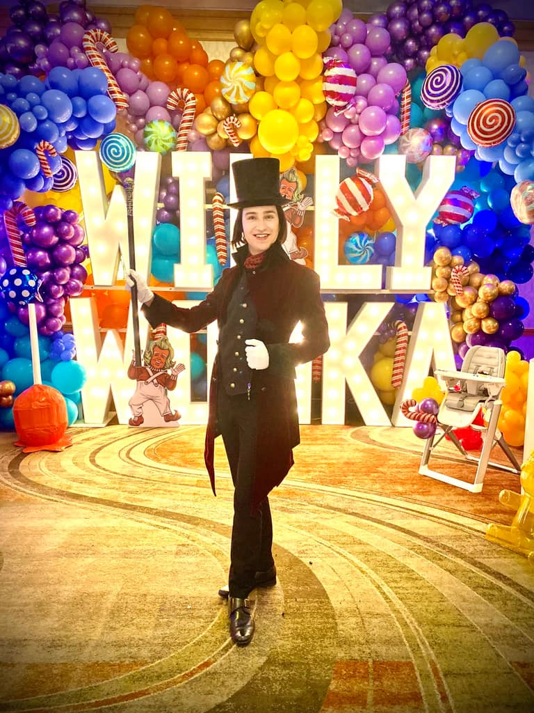 A Willy Wonka character before a candy-themed balloon arch and marquee letters